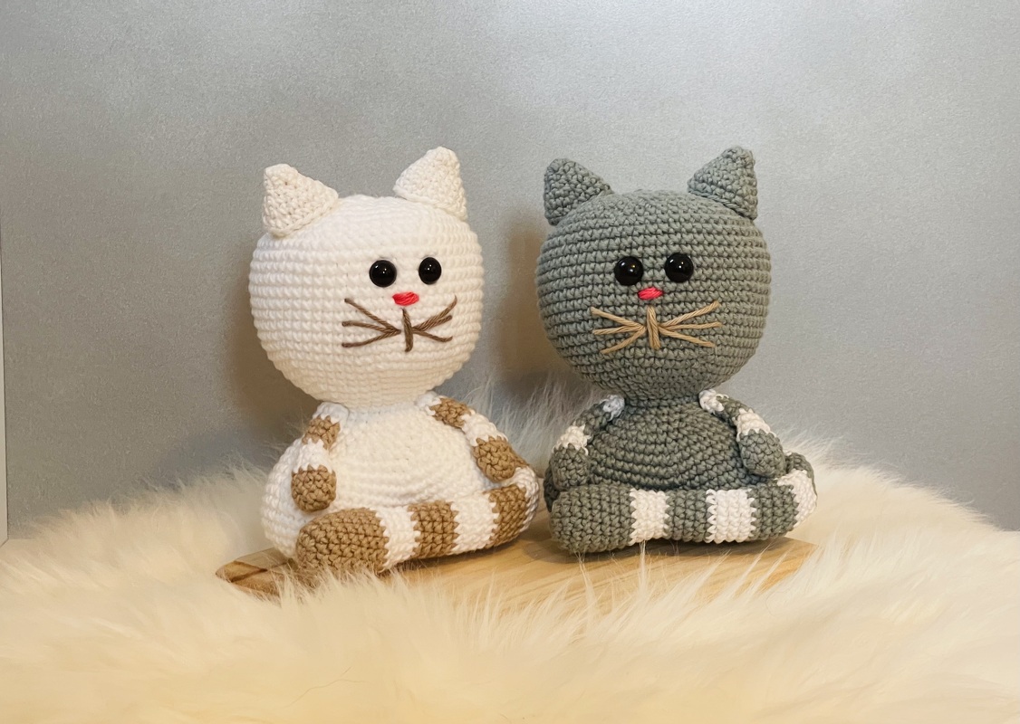 Zwei kompakte, schnell gehäkelte Amigurumi‑Katzen in creme‑braun und grau‑weiß auf hellem Untergrund