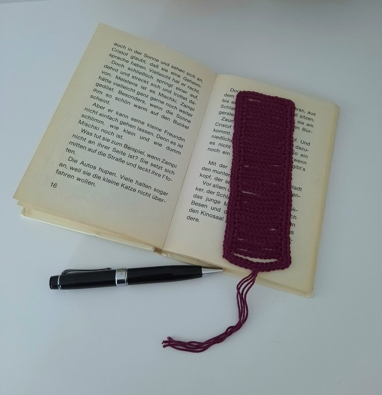 Gehäkeltes burgunderrotes Lesezeichen liegt in einem aufgeschlagenen Buch neben einem schwarzen Stift.