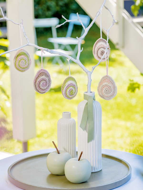 Gehäkelte Ostereier in Pastellfarben in Spiralform, hängend an einem weiß gestrichenen Ast, mit Vasen und Dekoäpfeln auf einem Tablett.