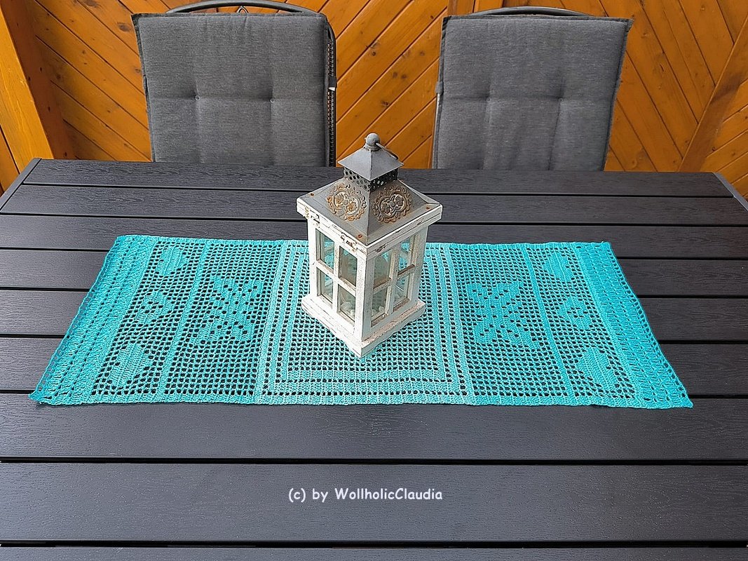 Gehäkelter türkiser Tischläufer mit Filetmuster und weißer Laterne auf dunklem Holztisch.
