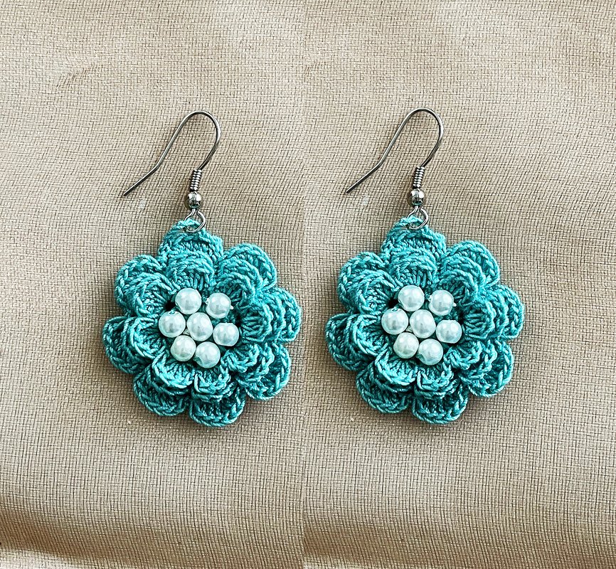 Pair of teal crocheted flower earrings with white pearl beads and silver-tone hooks on beige fabric.