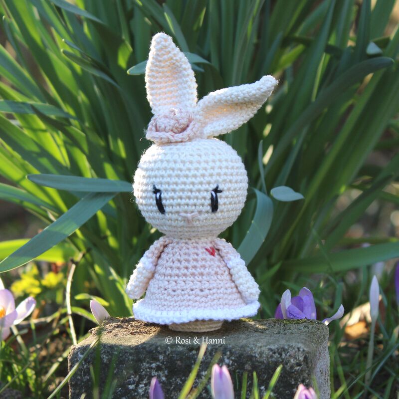 Gehäkelter Amigurumi-Hase im pastellrosa Kleid mit Blütenhaarband vor grünen Blättern und Krokussen