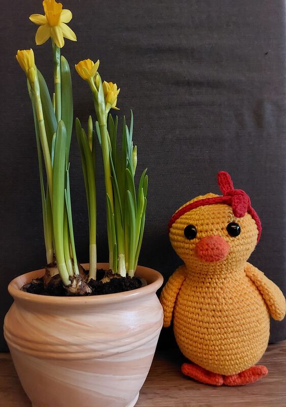 Handgehäkeltes Amigurumi-Küken mit roter Schleife neben Narzissen im Blumentopf