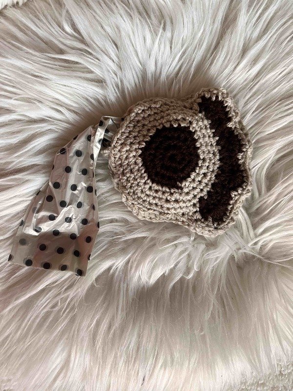 Gehäkelter Kotbeutelspender in Beige mit braunem Pfotenmotiv auf weißem Fellimitat; eine gepunktete Tüte ragt heraus.
