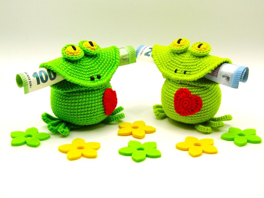 Zwei gehäkelte grüne Amigurumi-Frösche mit roten Herzen, gerollten Geldscheinen im Maul und bunten Filzblumen
