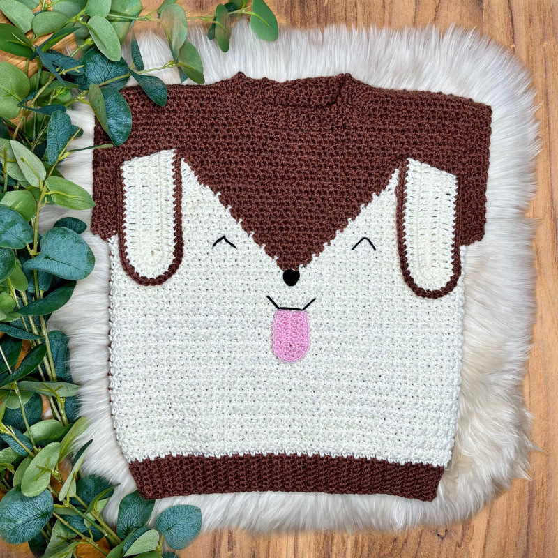 Gehäkelter braun-cremefarbener Kinderpullover mit Hundegesicht und rosa Zunge auf hellem Felluntergrund