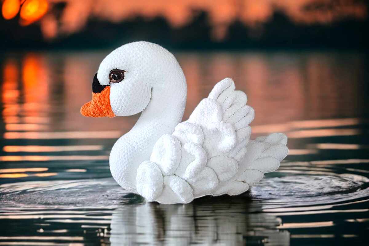 White crocheted swan amigurumi with orange beak floating on water at sunset.