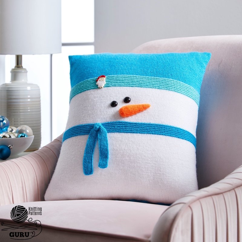 Square knitted snowman pillow with turquoise stripes, button eyes, orange carrot nose and small scarf on a pale armchair