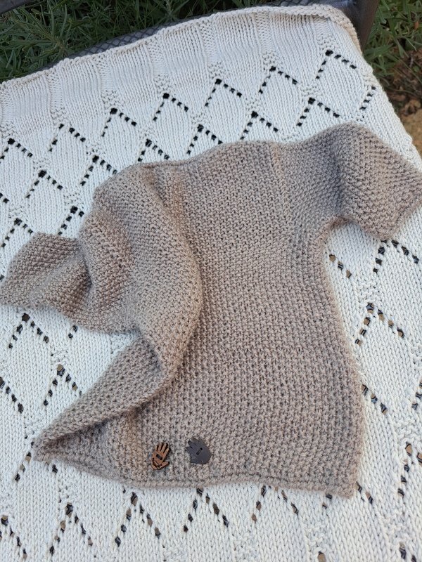 Gestrickter beiger Baby-Pullover mit kurzen Ärmeln auf weißer Lochmuster-Strickdecke, zwei Holzknöpfe am Saum