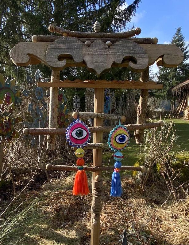Zwei gehäkelte Schutzaugen-Anhänger mit Perlen und Quasten an einem hölzernen Aufhänger im Garten