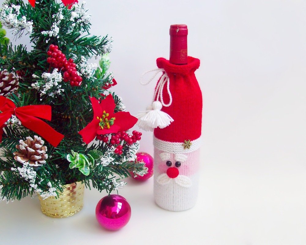 Red and white knitted Santa bottle cozy used as a holiday table accent beside a small decorated Christmas tree