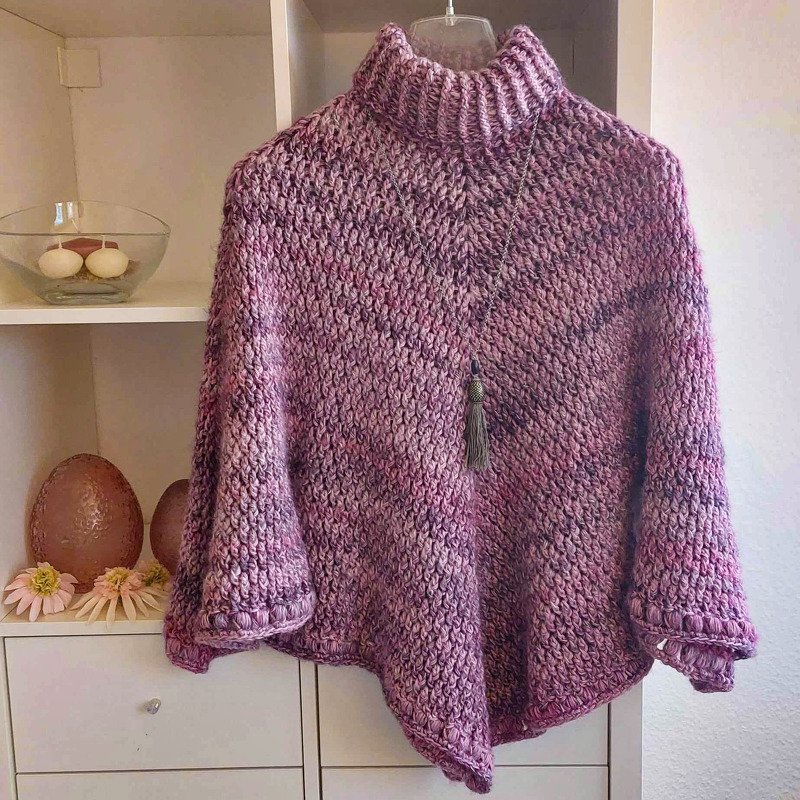 Gehäkelter Poncho in lila-melierten Tönen mit hohem geripptem Rollkragen an einem Kleiderbügel, mit Quastenanhänger.