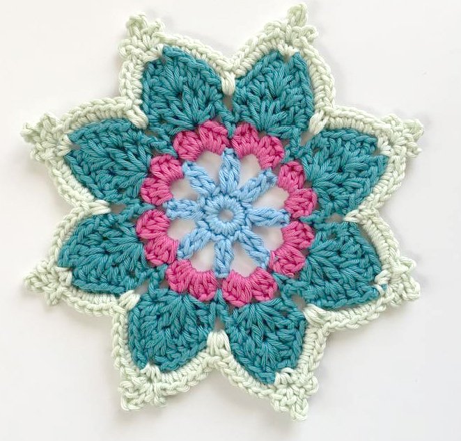 Nahaufnahme einer mehrlagigen, mehrfarbigen Häkel-Applikation in Blüten- oder Sternform mit Blau-, Pink- und Grüntönen.