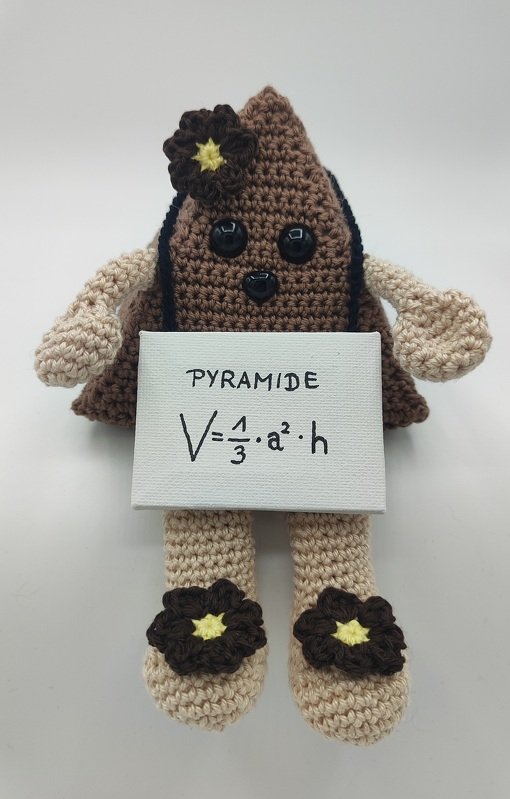 Pyramidenförmiges gehäkeltes Amigurumi in Braun und Beige mit Knopfaugen, Blumenapplikationen und Schild mit der Formel V = 1/3 · a² · h