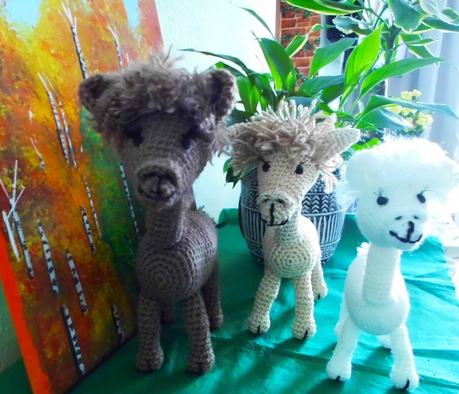 Drei gehäkelte Amigurumi-Lamas (braun, beige, weiß) auf grüner Stoffunterlage vor Pflanzen und herbstlicher Wanddekoration.