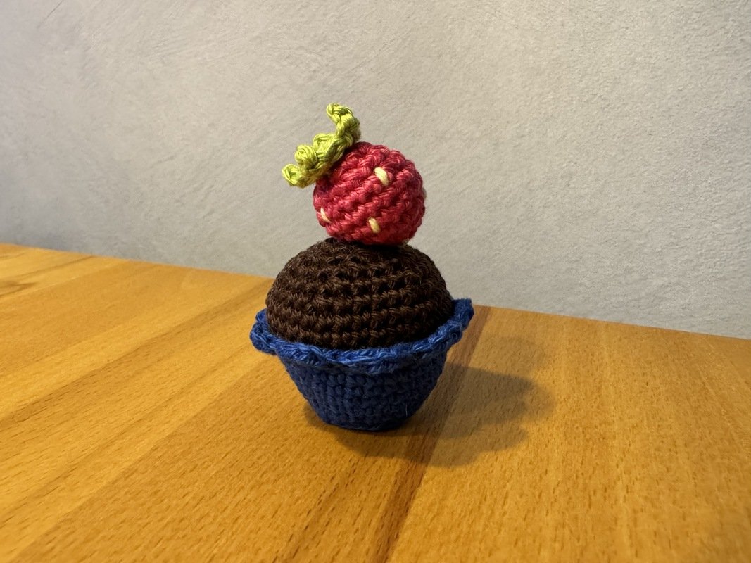 Gehäkelter Cupcake in blauem Förmchen mit brauner Kuppel und roter Erdbeere