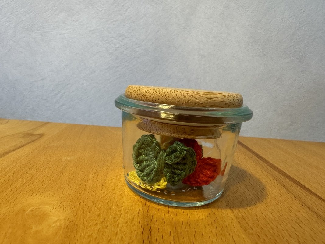 Kleine gehäkelte Herzen in Grün, Rot und Gelb in einem Glas auf einer Holzoberfläche