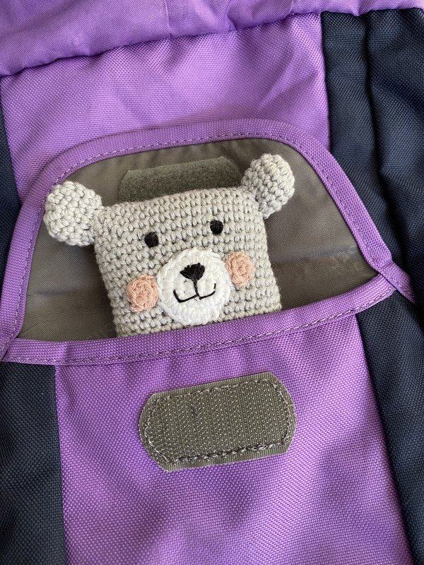 Kleiner gehäkelter Teddykopf aus grauem Garn in der Vordertasche eines lilafarbenen Rucksacks