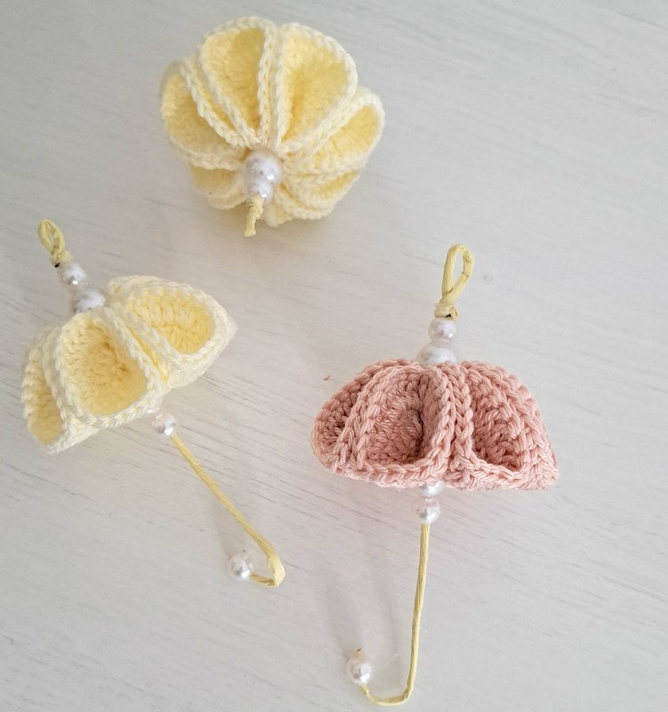 Zwei gehäkelte Mini‑Regenschirme in Hellgelb und Rosa und eine gehäkelte gelbe Blüte, alle mit kleinen Perlen verziert, auf hellem Untergrund.