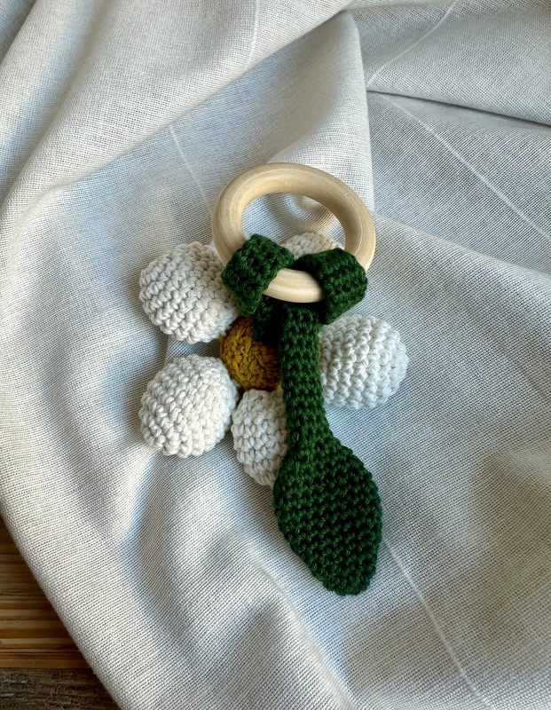Gehäkelter Baby-Beißring mit Holzring, weißen Häkelelementen, gelbem Zentrum und grünem Blatt auf hellem Stoff