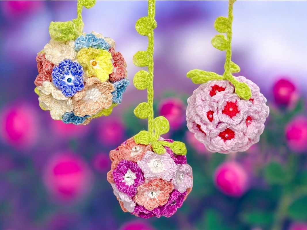 Three hanging crochet flower ball ornaments composed of many small multicolored flowers with green vine-like hangers against a blurred floral background.