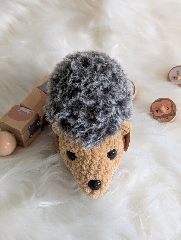 Gehäkelter Amigurumi-Igel mit beigem Gesicht und grauem, flauschigem Rücken, liegend auf hellem Untergrund