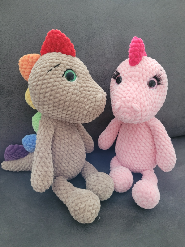 Zwei gehäkelte Amigurumi‑Drachen, beige mit Regenstacheln und rosa mit Wimpern, sitzend