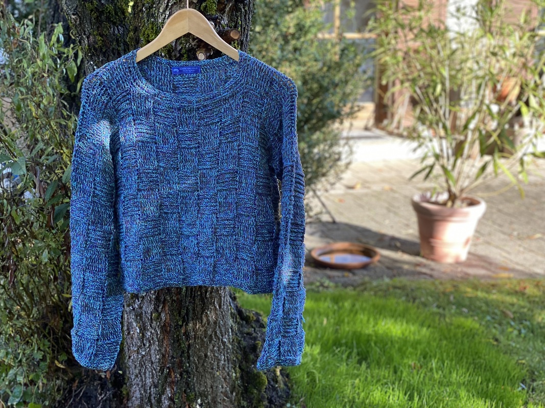 Variegated blue crocheted women's sweater with checkerboard texture hanging on a wooden hanger against a tree trunk