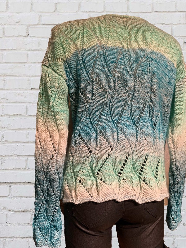 Nahaufnahme der Rückseite eines gestrickten Damenpullovers mit Blatt-Ajourmuster und Ombré-Farbverlauf in Creme, Blau und Grün vor weißer Ziegelwand.