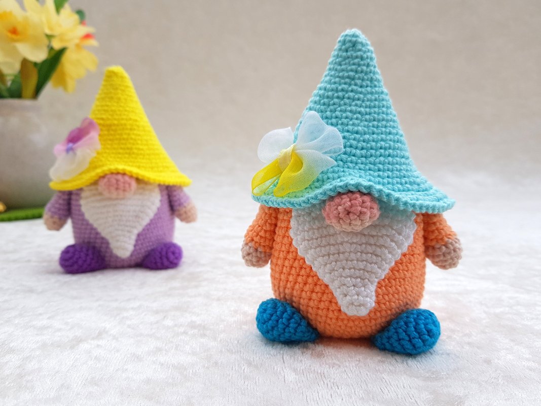 Zwei gehäkelte Amigurumi-Wichtel, vorne orange mit türkisfarbener Mütze und Blüte, im Hintergrund ein lila Wichtel mit gelber Mütze