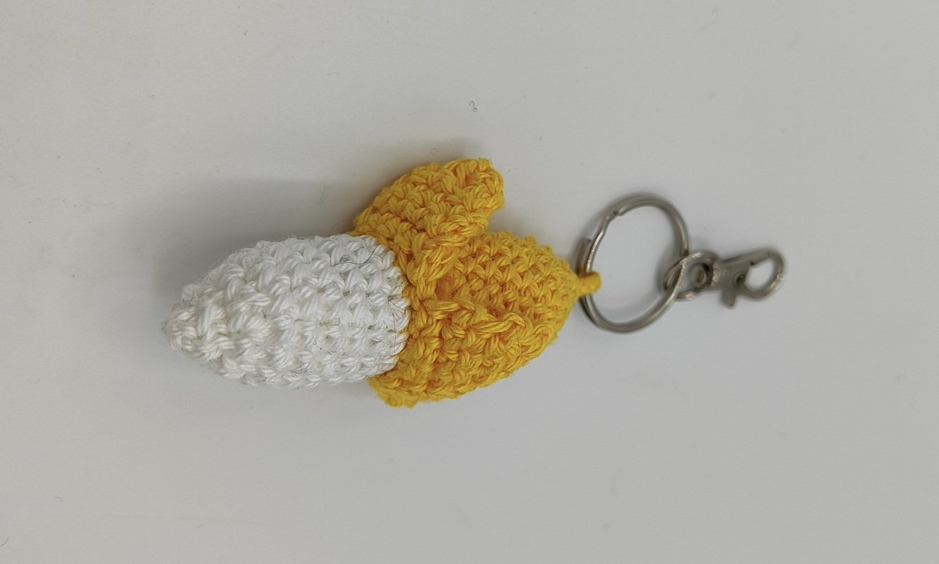 Gehäkelter Bananen‑Schlüsselanhänger in Gelb und Weiß mit Metallring und Karabiner auf hellem Hintergrund.