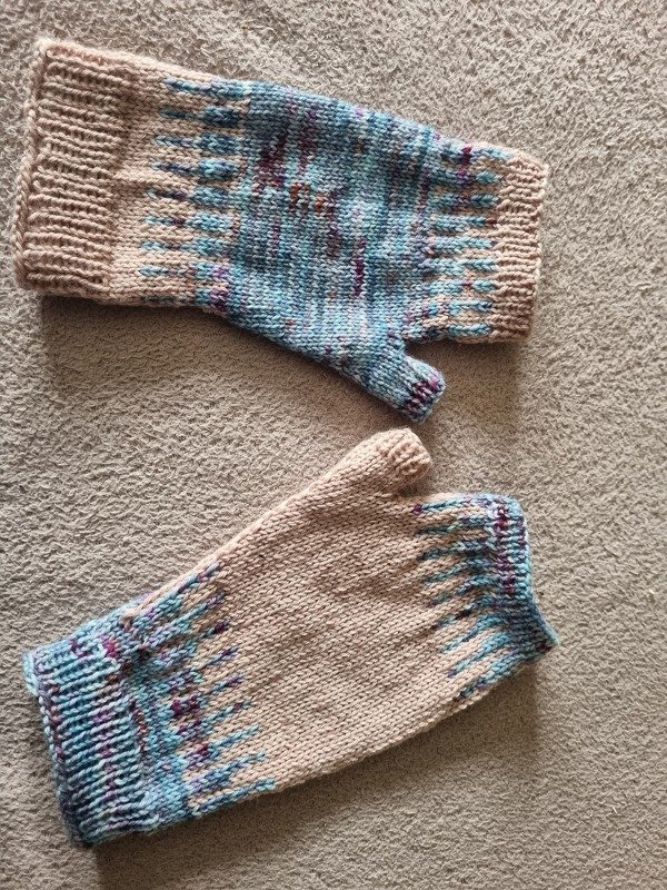 Paar gestrickter fingerloser Handschuhe in Beige mit blauen, türkisen und violetten Akzenten auf hellem Untergrund.
