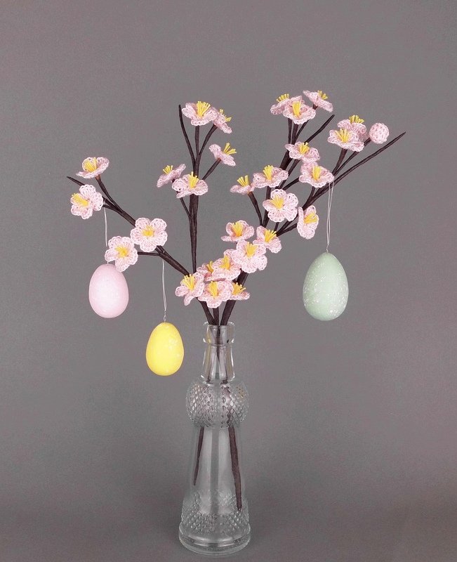 Clear glass vase with twig branches of pink crocheted flowers and three hanging pastel eggs (pink, yellow, mint) against a grey background