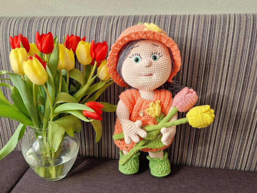 Gehäkelte Amigurumi-Puppe im orangefarbenen Outfit mit Häkeltulpen neben einer Vase mit Tulpen