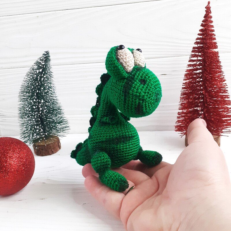 Small green crocheted dinosaur amigurumi with large eyes and back spikes sitting in an open hand, with miniature festive trees and a red bauble on a white surface.