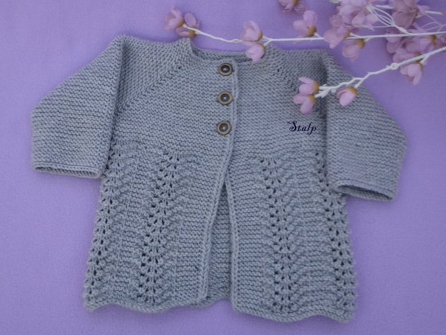 Graue, handgestrickte Babyjacke mit Lochmuster und drei Holzknöpfen, liegend auf lila Stoff mit zarten Blüten.