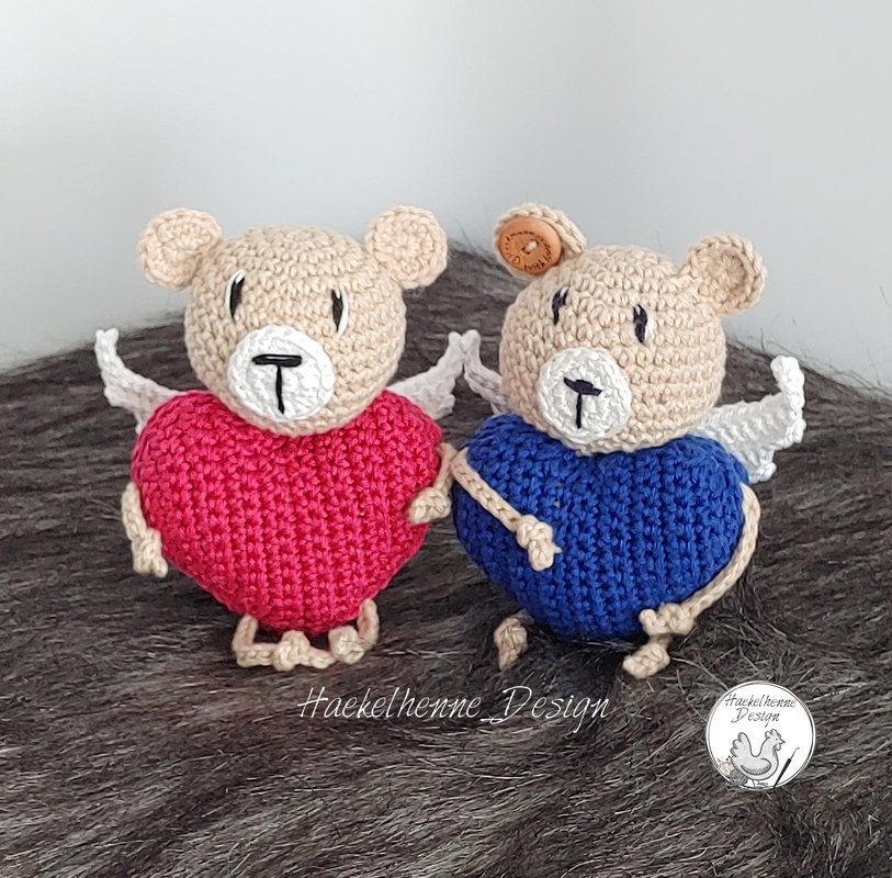 Zwei gehäkelte Amigurumi-Teddybären mit roten und blauen herzförmigen Körpern, beigen Köpfen und weißen Flügeln auf flauschigem Untergrund.