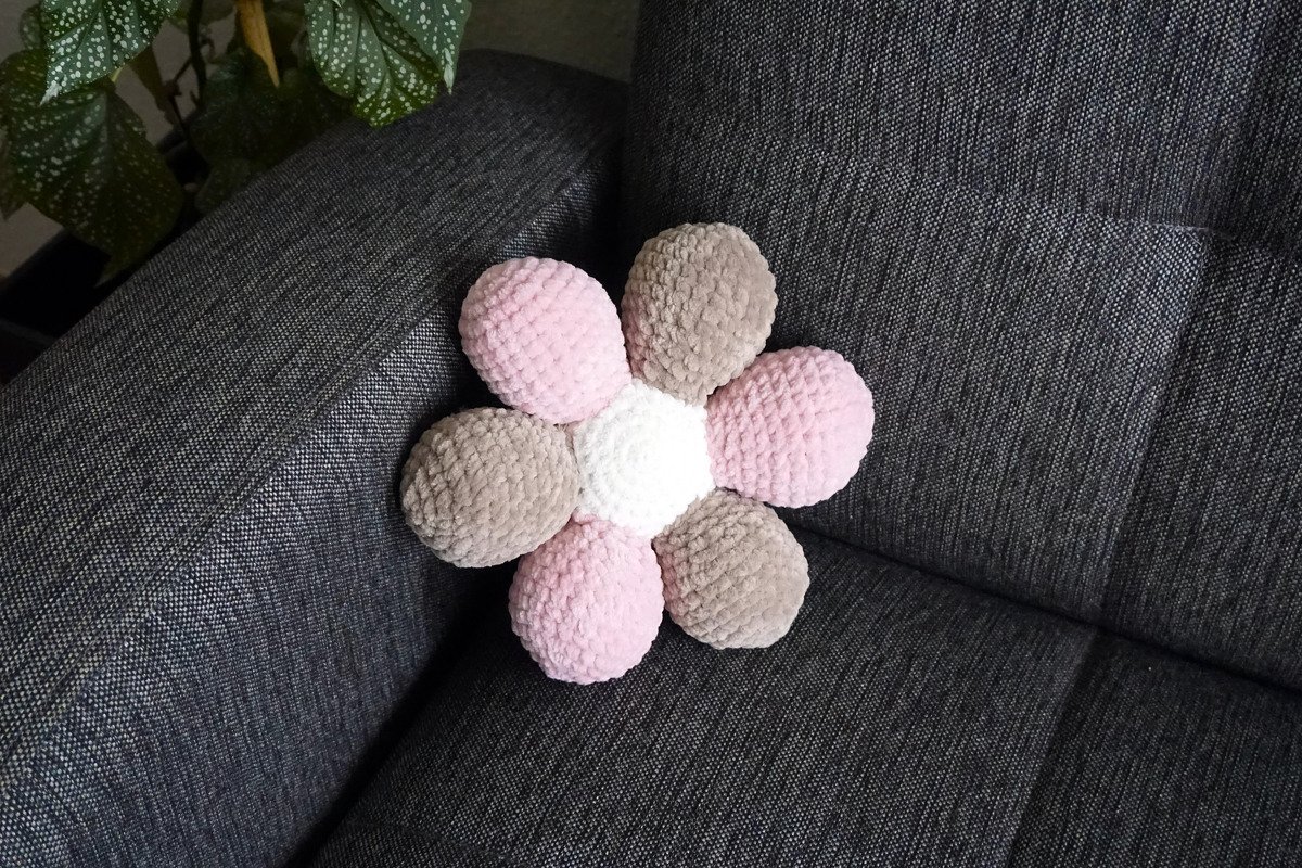 Gehäkeltes blütenförmiges Kissen in Rosa, Beige und Weiß auf dunkelgrauem Sofa.