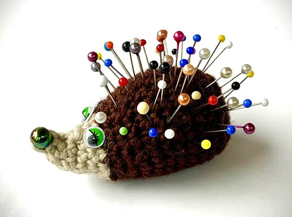 Gehäkeltes Igel-Nadelkissen mit grünen Augen und bunten Stecknadeln auf weißem Hintergrund