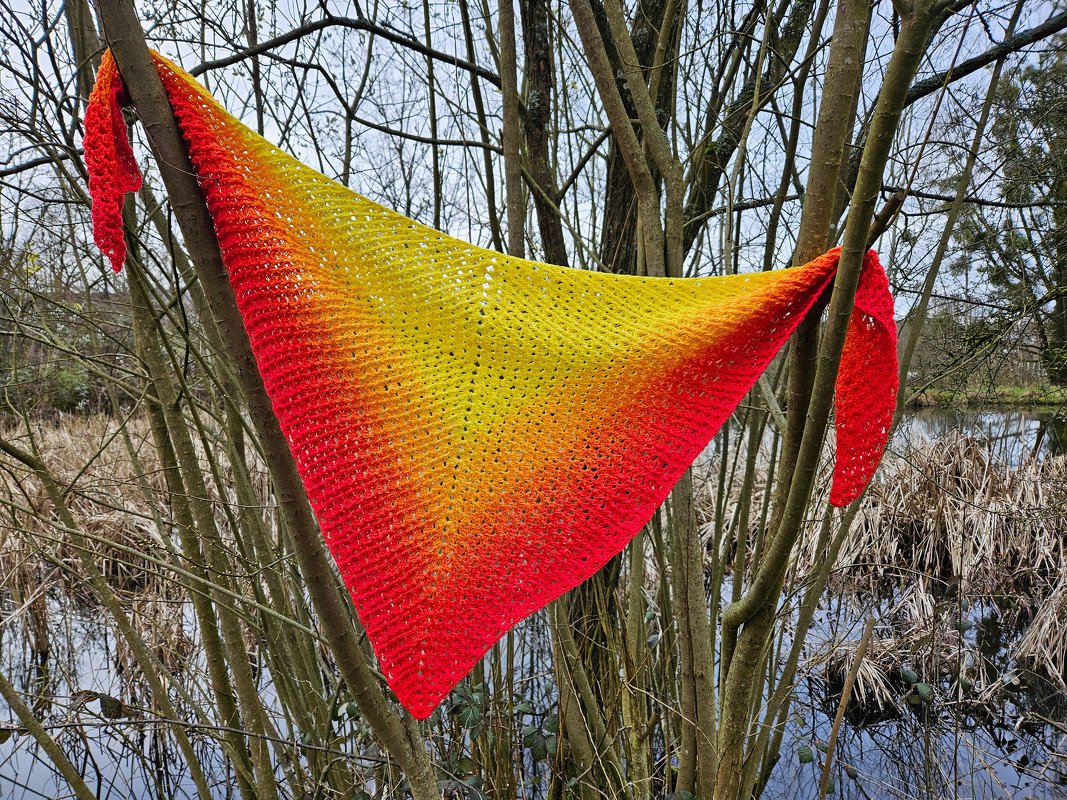 Gehäkeltes Dreieckstuch mit Farbverlauf von Gelb zu Rot, aufgehängt an Baumzweigen vor einem Teich