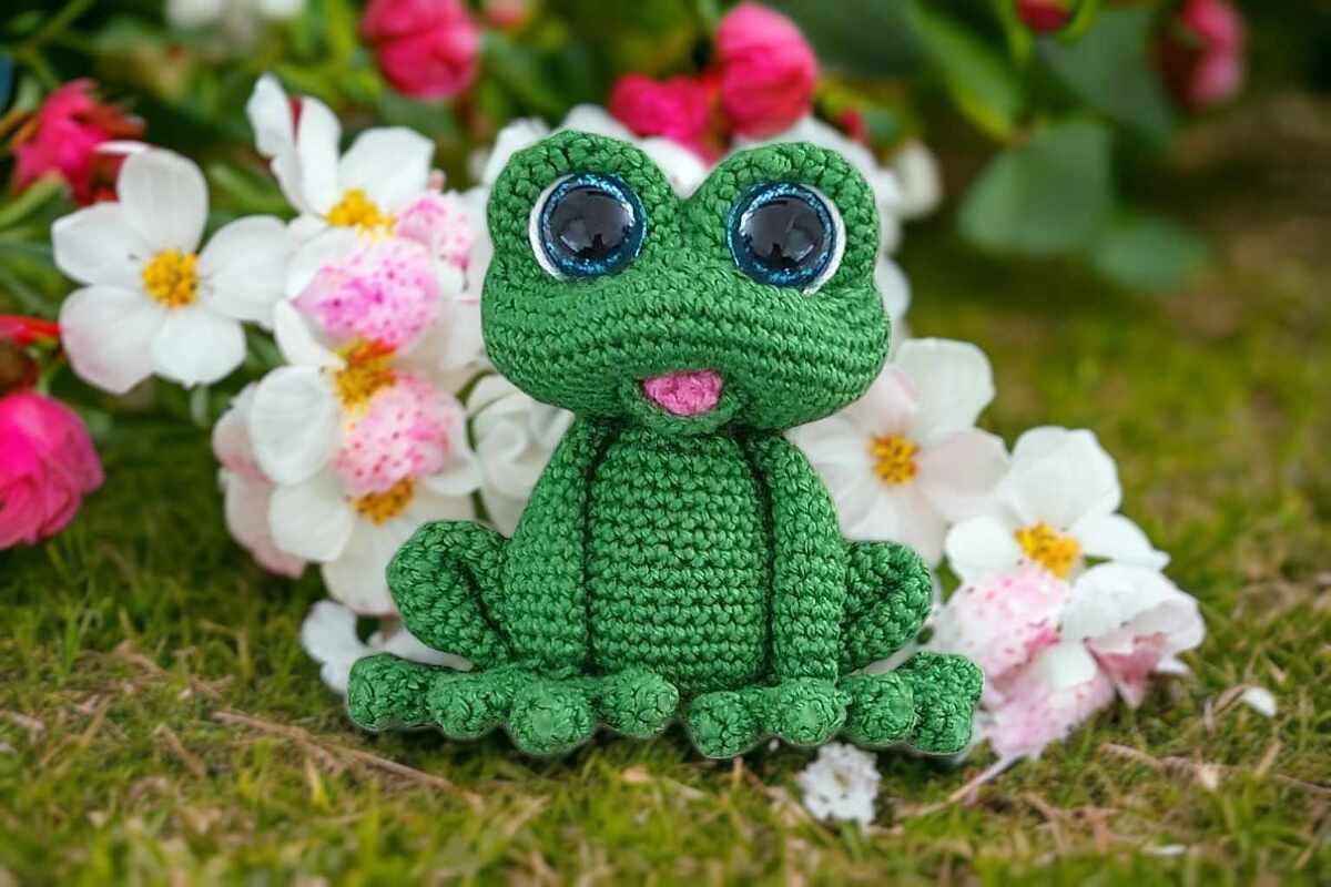 Green crocheted frog amigurumi with large blue eyes and pink tongue on grass with flowers.