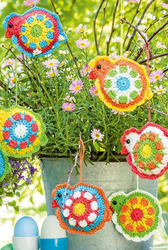 Bunte, rund gehäkelte Vogelanhänger hängen an Zweigen neben Ostereiern.