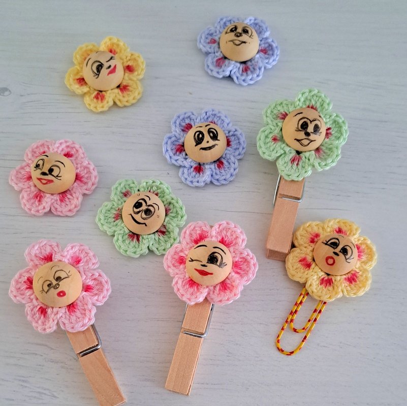 Mehrere kleine gehäkelte Blüten in Pastellfarben mit Holzknöpfen, an Wäsche- und Büroklammern befestigt