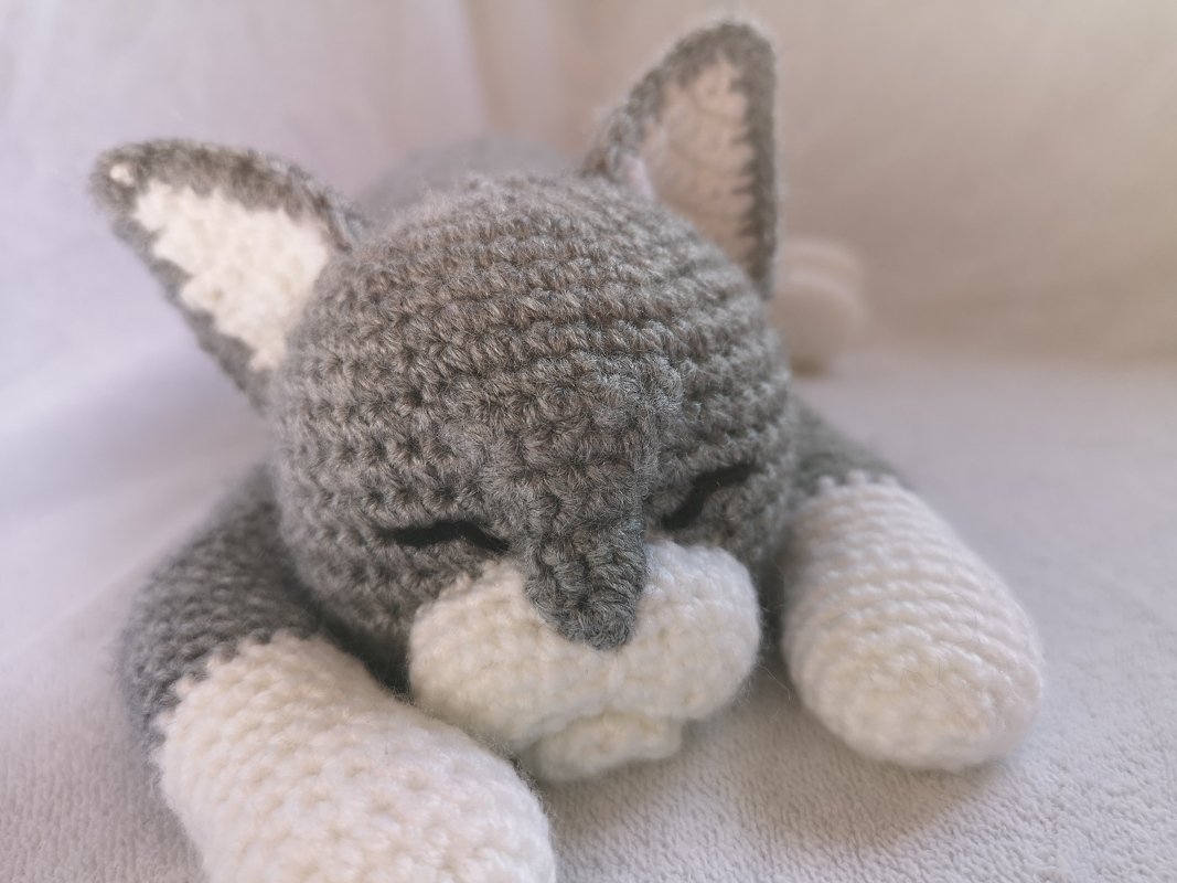 Crocheted grey and white amigurumi cat toy sleeping on a light surface, close-up.