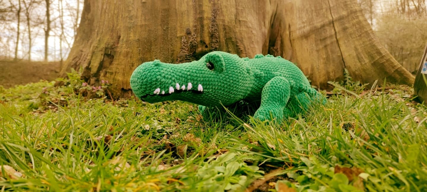 Grünes gehäkeltes Krokodil-Amigurumi mit weißen Zähnen im Gras vor einem Baumstamm.