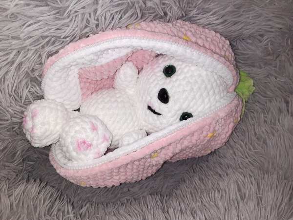 Weißer gehäkelter Amigurumi-Hase mit rosa Akzenten in geöffneter rosa Erdbeertasche auf grauem Flauschuntergrund