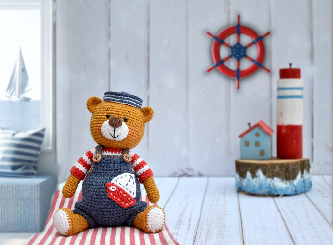 Brown crocheted teddy bear wearing blue overalls with a small boat appliqué, a red-and-white striped shirt, and a blue sailor hat, seated on a striped blanket with nautical decor in the background.