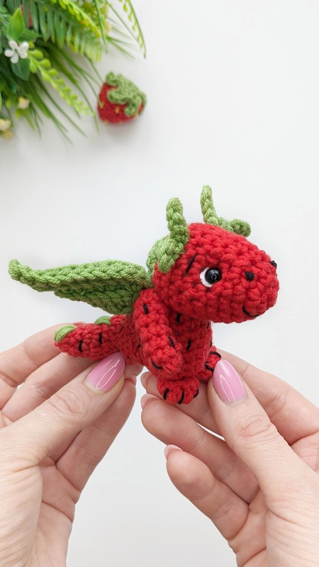 Hands holding a small red crocheted strawberry dragon with green wings and black seed-like stitches.