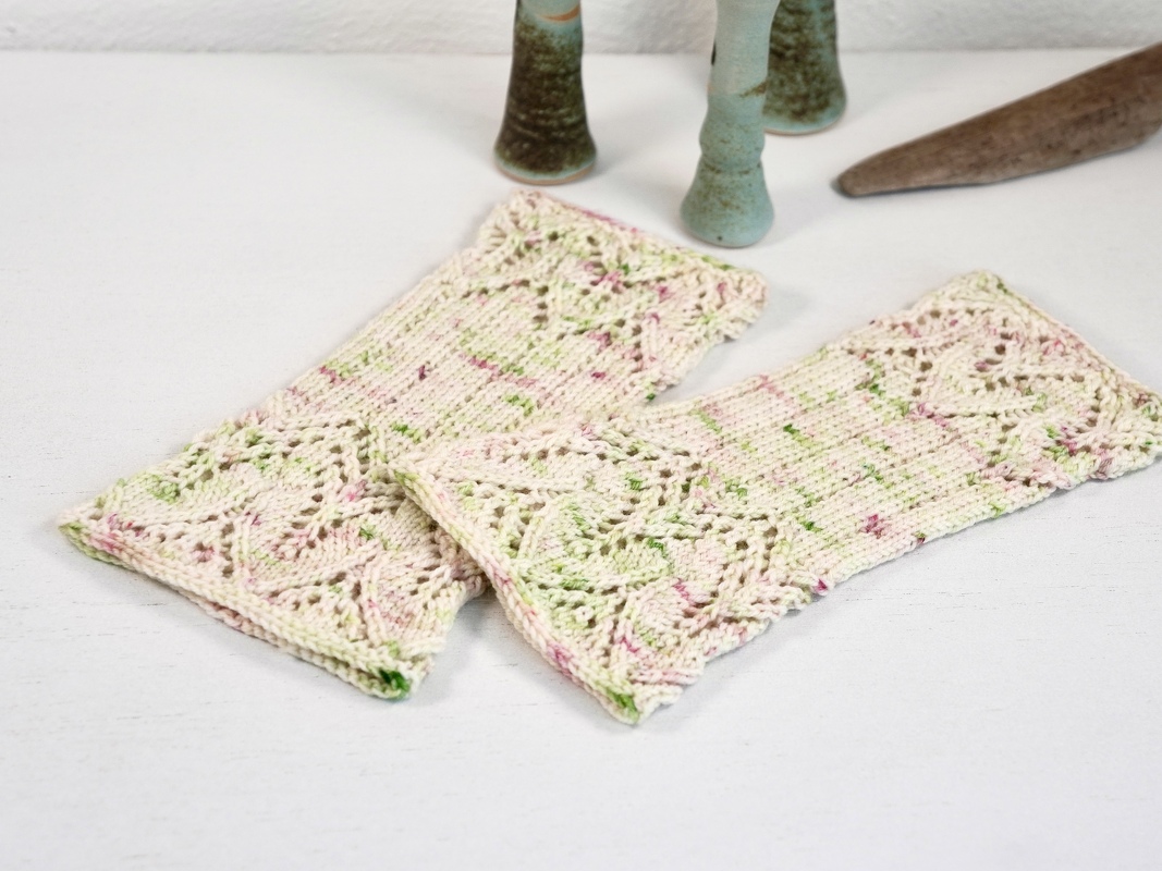 Gestrickte, fingerlose Armstulpen in Creme mit rosa- und grünen Sprenkeln und durchbrochenem Muster auf weißer Fläche.