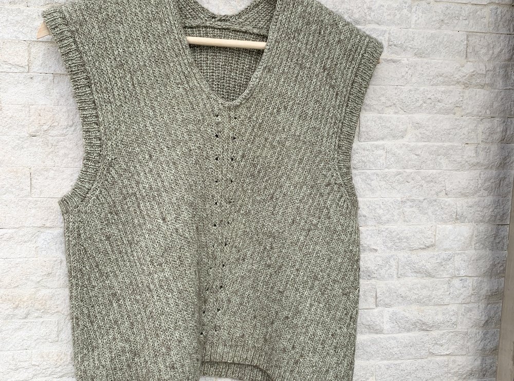 Gestrickte ärmellose Damenweste in Grüngrau-Melange mit V-Ausschnitt, aufgehängt an einem Holzbügel vor heller Steinwand.